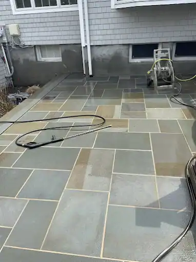 A patio made of large rectangular gray stone tiles is being power washed in Nassau & Suffolk County, NY. A pressure washer is visible at the far end, with some tiles wet and others dry, highlighting the contrast in color.