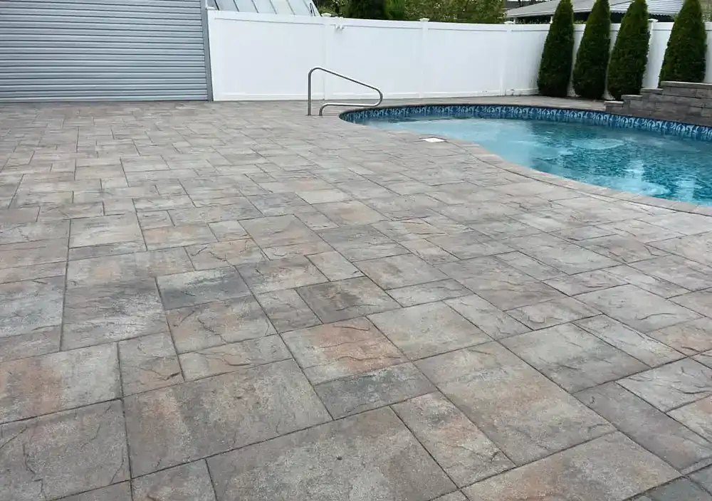 A backyard with a swimming pool featuring a blue patterned edge, surrounded by gray stone pavers freshly cleaned by power washing Nassau & Suffolk County, NY, a white fence, tall shrubs, and a closed metal roller door in the background.