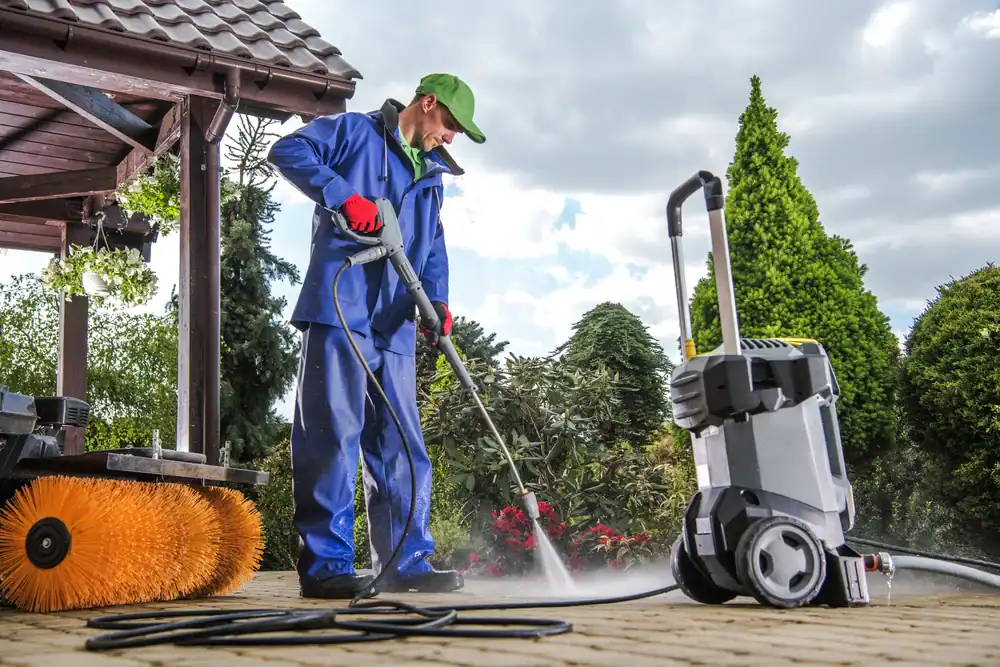 A person in blue work clothes and a green cap uses a pressure washer to clean an outdoor patio in NY. Nearby is a large broom attachment, with trees and bushes in the background—power washing Nassau & Suffolk County done right.